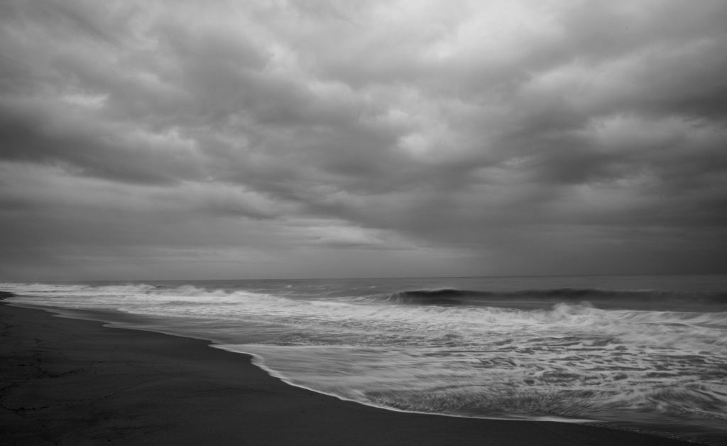 Ondas avançam sobre a faixa de areia sob um céu encoberto e cinzento.