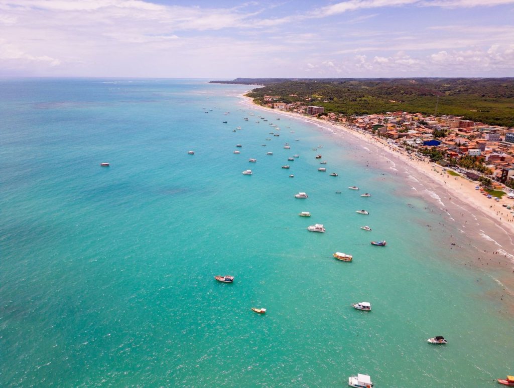 Vista aérea de uma praia brasileira de águas azul-turquesa, com diversas embarcações próximas à costa.